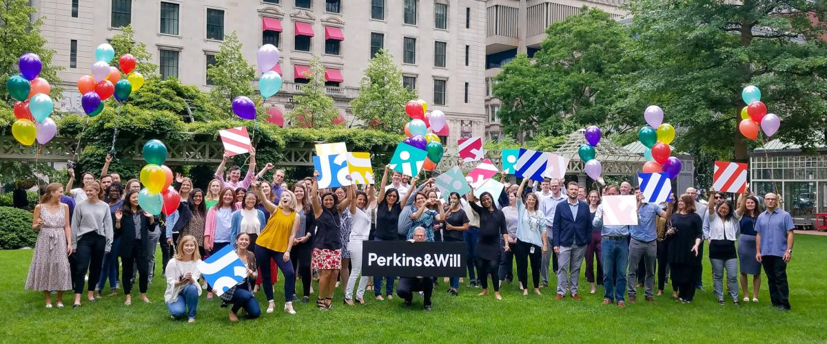 Employees gathered in a park with balloons and the Perkins&Will logo on a sign