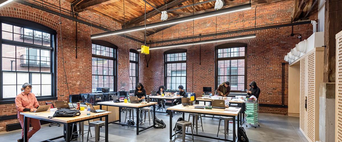 Photograph of a room with brick walls and wood beam ceiling. Six people work on electronics at industrial tables.