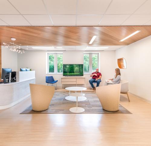 Photo of a well-lit healthcare center lobby with people seated in the waiting area and behind a check-in desk. Interior materials consist of light woods and organic forms that prioritize ample natural light.